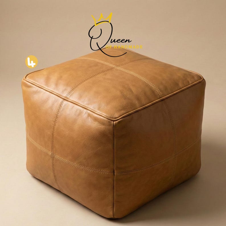 May include: A square, light brown leather ottoman with stitched detailing. The ottoman has a cube shape and is placed on a neutral background. The text "Queen of Handmade" is visible in the upper right corner.