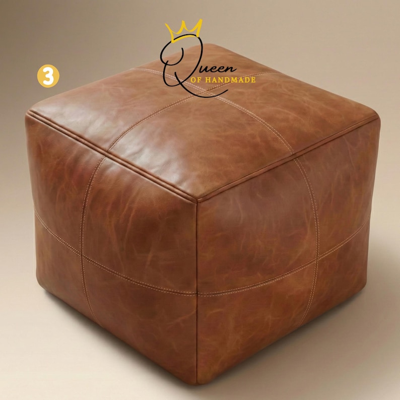 May include: A square, brown leather ottoman with stitched detailing. The ottoman has a cube shape and is made of a rich, textured leather. The text "Queen of Handmade" is visible on the top right corner.
