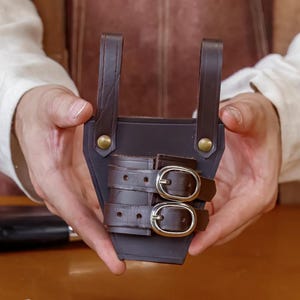 May include: Two leather sheaths, one black and one brown, are displayed. The brown sheath is held up, showcasing two buckles. The black sheath, with two buckles, rests on a wooden surface. Both sheaths have straps and rivets, suggesting a handcrafted design.
