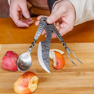 May include: A set of medieval-style cutlery, including a spoon, knife, and fork, held over a wooden cutting board. The utensils have twisted handles. Sliced apples and a whole red potato are on the board. A black pan is in the background.