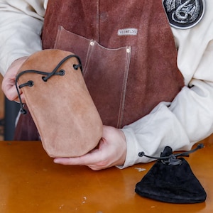May include: A brown leather pouch with a drawstring closure and black leather straps. The pouch is being held by a person wearing a brown leather apron with a dragon emblem.