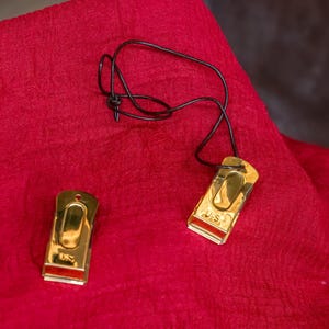 Pu&ograve; includere: Due fermasoldi in metallo color oro con un cordino nero, appoggiati su un tessuto rosso. Le clip hanno una forma rettangolare con la parte superiore curva e una striscia rossa nella parte inferiore. Lo sfondo &egrave; un tessuto rosso.