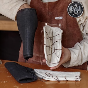 May include: A blacksmith's workshop scene featuring protective arm guards and gloves. The arm guards are black, and the gloves are black. One arm guard is held up, and the other items are on a wooden surface. A brown leather apron is worn.