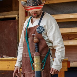 May include: A brown leather apron with a sword sheath and a decorative badge. The sword has a wooden handle and a brass guard. The person is wearing a cowboy hat with a stars and stripes design.