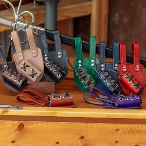 May include: A collection of colorful leather sword scabbards in various colors, including brown, tan, black, green, blue, red, and purple. Each scabbard features a laced design and is displayed on a wooden surface.