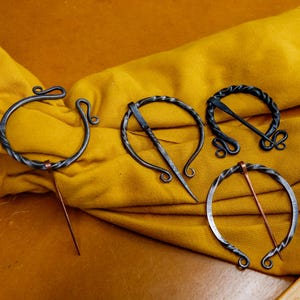 May include: Four handcrafted metal shawl pins with unique designs. The pins are displayed on a yellow textile. Each pin features a circular or oval shape with decorative elements and a pin for securing the shawl.