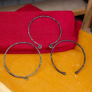 May include: Three silver-toned metal torc necklaces with textured designs. One has a hook closure, and two have spiral or curled ends. They rest on a yellow surface with a red fabric in the background.