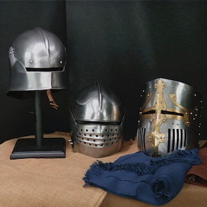 May include: Three medieval helmets are displayed on a burlap surface against a dark background. The helmets are made of metal and feature different designs, including a helmet on a stand, a helmet with a visor, and a helmet with gold accents.