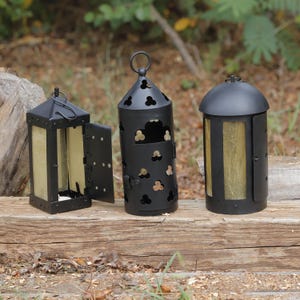 May include: Three black metal lanterns with glass panels. The left lantern is square with an open door. The center lantern is cylindrical with clover-shaped cutouts and a top ring. The right lantern is cylindrical with a domed top.