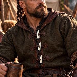 May include: A dark olive green hooded tunic featuring three white toggle closures with brown leather loops down the center front. The garment has short sleeves with intricate braided trim at the cuffs. The text MEDIEVAL CLOTHING is visible.