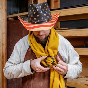 May include: A person wearing a straw cowboy hat with a red, white, and blue American flag design. The person is wearing a yellow scarf and a brown leather apron. A black metal clasp is being attached to the scarf.