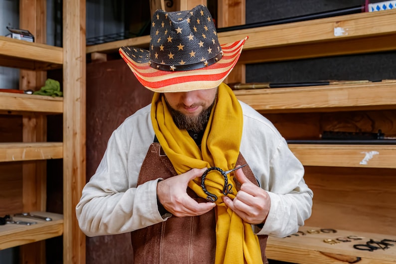 May include: A person wearing a straw cowboy hat with a red, white, and blue American flag design. The person is wearing a yellow scarf and a brown leather apron. A black metal clasp is being attached to the scarf.