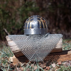 May include: A medieval helmet with a silver base, black and gold detailing, and a riveted pattern. It sits atop a chainmail coif, displayed on a weathered wooden log, with a blurred natural backdrop.
