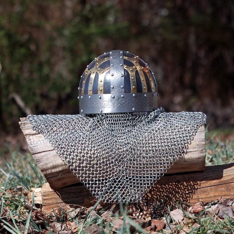 May include: A medieval helmet with a silver base, black and gold detailing, and a riveted pattern. It sits atop a chainmail coif, displayed on a weathered wooden log, with a blurred natural backdrop.