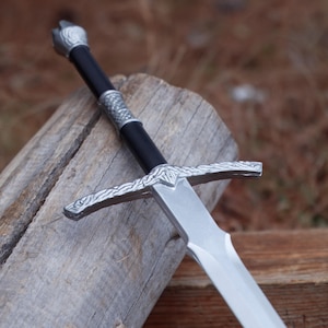 May include: A silver and black sword with a crossguard and a detailed handle. The blade is silver and the handle is black with silver accents. The sword is resting on a piece of wood.