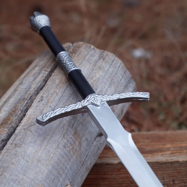 May include: A silver and black sword with a crossguard and a detailed handle. The blade is silver and the handle is black with silver accents. The sword is resting on a piece of wood.