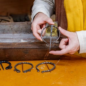 May include: A person holds a silver metal brooch with a long pin, showcasing its design. Three other brooches are displayed on a yellow surface. The brooches have a twisted metal design and are likely handmade.