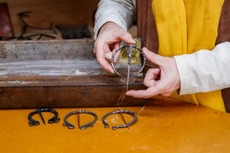 May include: A person holds a silver metal brooch with a long pin, showcasing its design. Three other brooches are displayed on a yellow surface. The brooches have a twisted metal design and are likely handmade.