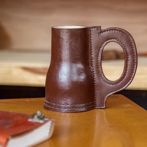 May include: A brown leather mug with a distinctive handle. The mug has a cylindrical body and a stitched handle. The interior is white. The mug is on a wooden surface, with a book in the foreground.