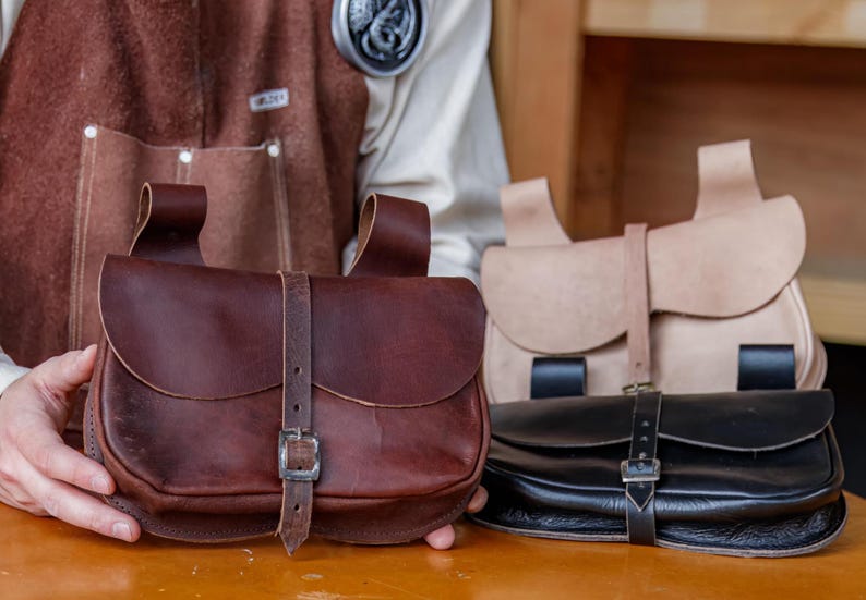 Op de afbeelding: Drie leren tassen met gespen en banden. De tassen zijn bruin, beige en zwart.
