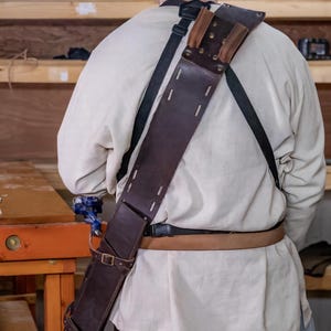 May include: A brown leather scabbard with a strap and buckle. The scabbard is worn over the shoulder and secured at the waist with a belt.