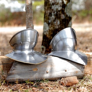 May include: Two polished steel gorgets, neck and shoulder armor, rest on a weathered wooden surface. The armor has a curved design with rivets and a reflective surface, suggesting historical or fantasy themes.