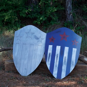 May include: Two wooden shields, one silver and one blue, are displayed outdoors. The silver shield has three stars and vertical stripes. The blue shield features three red stars and white stripes, evoking a patriotic theme.