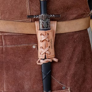 May include: A medieval sword with a black handle and silver crossguard, in a tan leather scabbard. The scabbard is laced with leather and has the text "USACraftmen". The sword is attached to a brown leather belt, typical of historical weaponry.