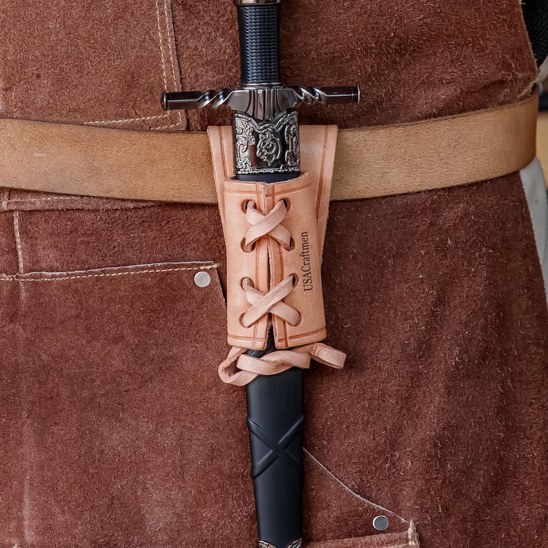 May include: A medieval sword with a black handle and silver crossguard, in a tan leather scabbard. The scabbard is laced with leather and has the text "USACraftmen". The sword is attached to a brown leather belt, typical of historical weaponry.