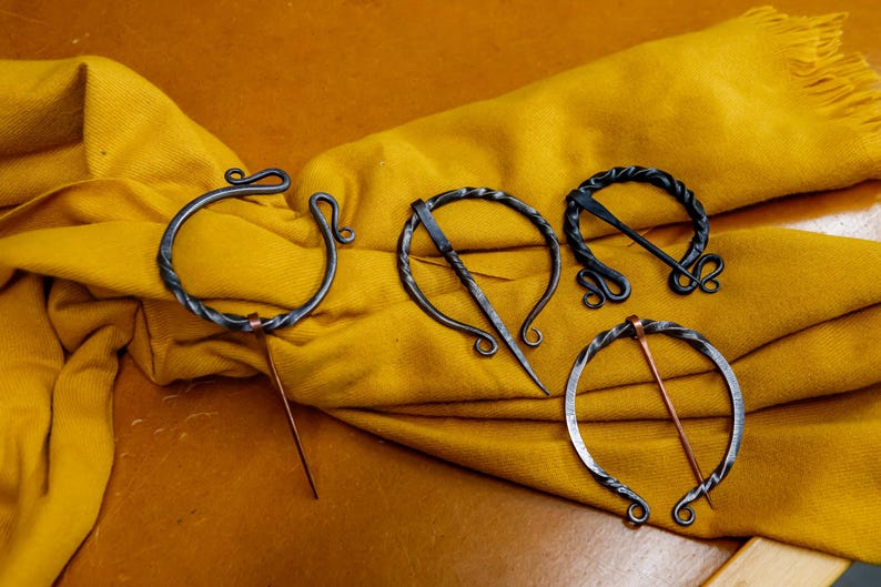 May include: Four handcrafted metal shawl pins with unique designs. The pins are displayed on a yellow textile. Each pin features a circular or oval shape with decorative elements and a pin for securing the shawl.