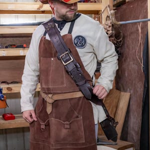 May include: A brown leather apron with a large pocket on the front and a thick leather strap that goes over the shoulder. The apron is worn by a person wearing a white shirt with a patch on the chest.