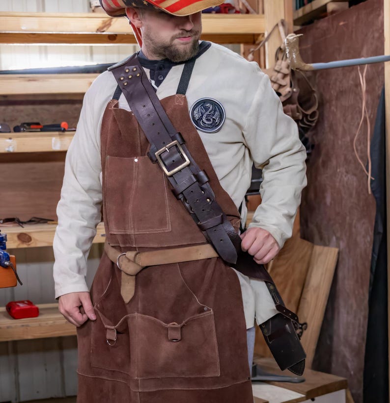 May include: A brown leather apron with a large pocket on the front and a thick leather strap that goes over the shoulder. The apron is worn by a person wearing a white shirt with a patch on the chest.