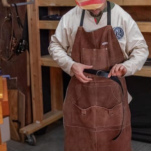 May include: A brown leather apron with two large pockets and a black leather belt. The apron is being worn by a person with a white shirt and a red and white striped hat.