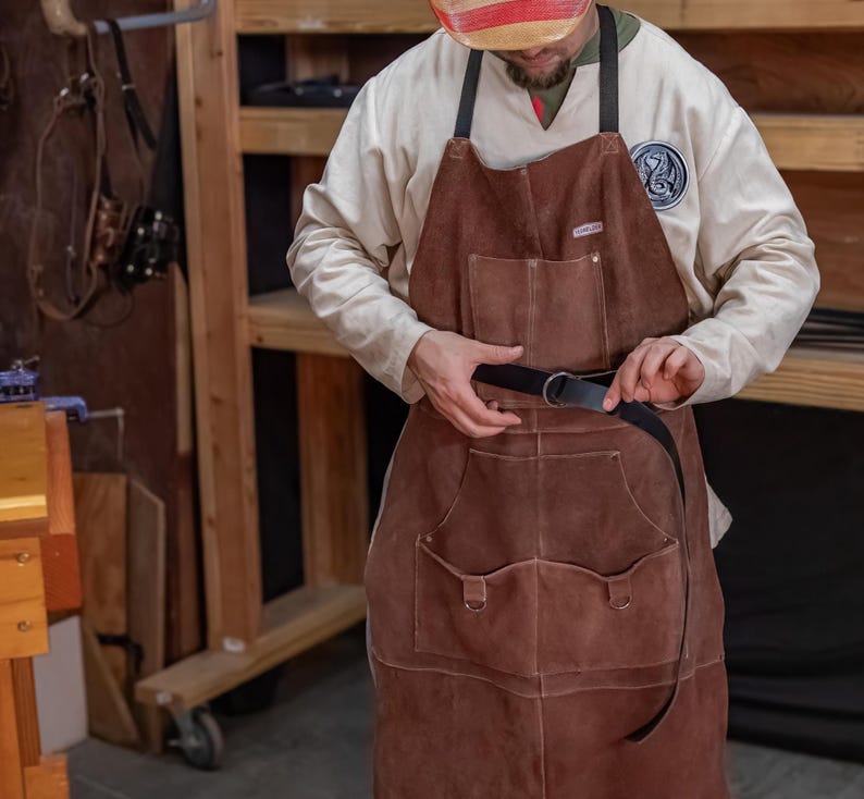 May include: A brown leather apron with two large pockets and a black leather belt. The apron is being worn by a person with a white shirt and a red and white striped hat.