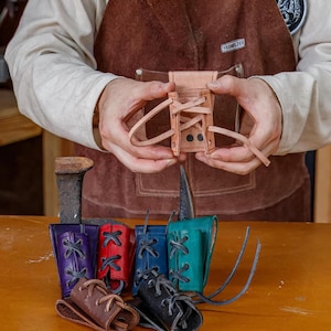 May include: A small, light brown leather pouch with a laced closure is held in the hands. Below, several colourful leather pouches in purple, red, blue, teal, brown, and black are displayed, each with a laced design.