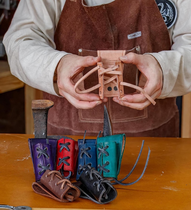 May include: A small, light brown leather pouch with a laced closure is held in the hands. Below, several colourful leather pouches in purple, red, blue, teal, brown, and black are displayed, each with a laced design.