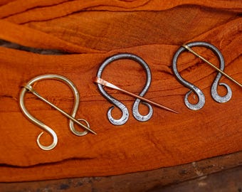 Op de afbeelding: Drie metalen mantelspelden met een kenmerkend ontwerp, elk met een gebogen lichaam en een rechte speld. Eén is goudkleurig, de andere zijn zilverkleurig met koperen en gouden spelden. Ze rusten op een oranje stoffen achtergrond.