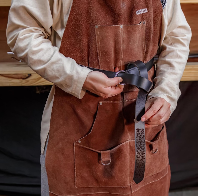 May include: A person wearing a brown leather apron with a black leather belt with a Celtic knot design. The belt has a silver buckle and is fastened with a ring.