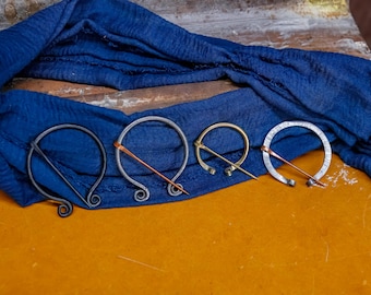 May include: Four metal brooches with pins rest on a blue fabric and a yellow-orange surface. The brooches vary in size and design, with circular and oval shapes. Some have copper-colored accents, and the metal finishes range from dark to silver.