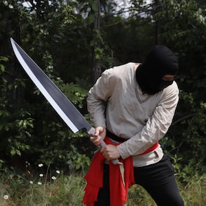 May include: A large, black and silver sword with a wide blade. The sword is held by a person wearing a black balaclava, a light-colored shirt, and black pants. A red sash is wrapped around the waist.