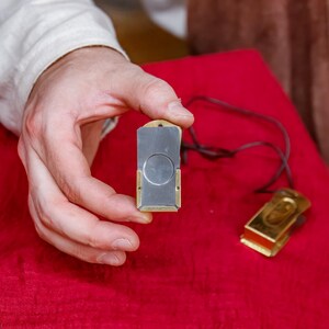 Pu&ograve; includere: Un tagliasigari rettangolare con una lama argentata e dettagli color oro &egrave; tenuto sopra una superficie rossa. &Egrave; visibile anche un secondo tagliasigari con finitura dorata e un cordino nero.
