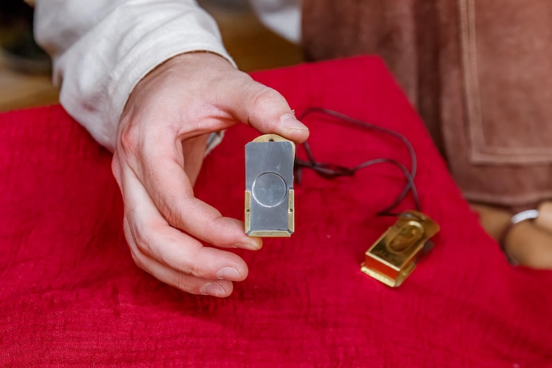 Pu&ograve; includere: Un tagliasigari rettangolare con una lama argentata e dettagli color oro &egrave; tenuto sopra una superficie rossa. &Egrave; visibile anche un secondo tagliasigari con finitura dorata e un cordino nero.