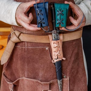 May include: A medieval dagger with a black handle and silver detailing, presented with a tan leather sheath and two small leather pouches in blue and teal. The sheath and pouches feature decorative lacing.