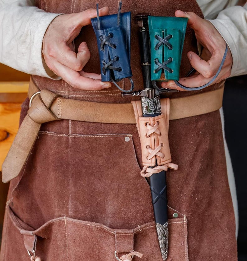May include: A medieval dagger with a black handle and silver detailing, presented with a tan leather sheath and two small leather pouches in blue and teal. The sheath and pouches feature decorative lacing.