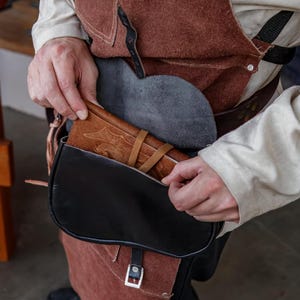 Op de afbeelding: Een persoon reikt in een zwarte leren tas met een bruine leren klep. De tas wordt gedragen aan een bruine leren riem. De tas is open en een bruin leren dagboek is zichtbaar in de tas.