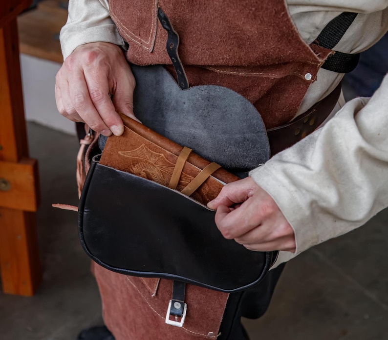 Op de afbeelding: Een persoon reikt in een zwarte leren tas met een bruine leren klep. De tas wordt gedragen aan een bruine leren riem. De tas is open en een bruin leren dagboek is zichtbaar in de tas.