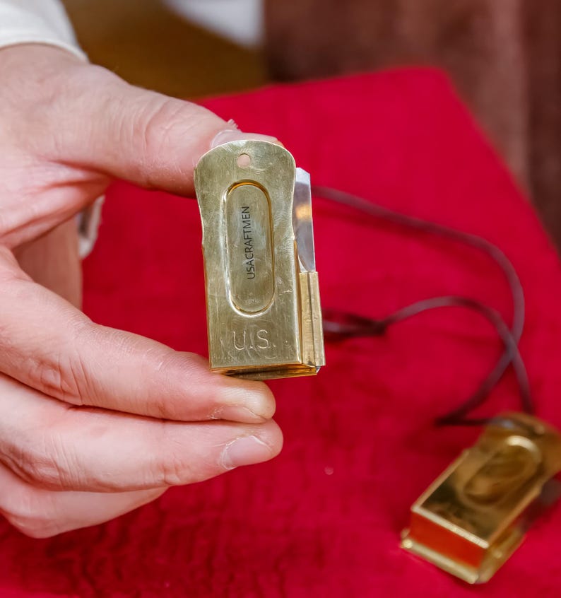 Pu&ograve; includere: Un coltello tascabile rettangolare color oro con la scritta "USA CRAFTMEN" e "U.S." incisa sul davanti. Il coltello ha una piccola lama e un cordino marrone scuro. Un altro coltello &egrave; visibile sullo sfondo.