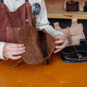 Op de afbeelding: Een bruine leren tas met een klep is open om de binnenkant te laten zien. De tas is gemaakt van zacht, soepel leer en heeft een eenvoudig, klassiek ontwerp.