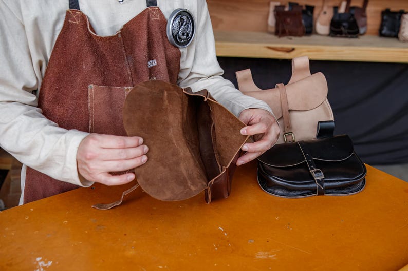 Op de afbeelding: Een bruine leren tas met een klep is open om de binnenkant te laten zien. De tas is gemaakt van zacht, soepel leer en heeft een eenvoudig, klassiek ontwerp.