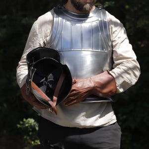 Pu&ograve; includere: Una corazza in acciaio lucido indossata sopra una camicia beige a maniche lunghe, abbinata a guanti in pelle marrone. Un elmo in metallo nero con visiera aperta e sottogola in pelle marrone &egrave; tenuto in mano, mostrando l'imbottitura interna.